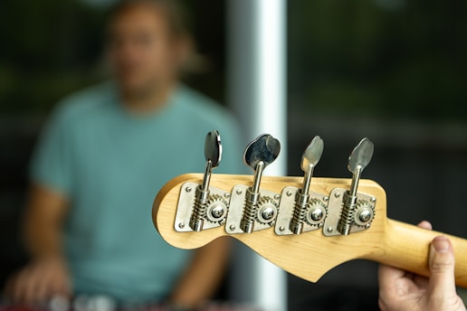 A close-up view of a bass guitar headstock with tuning pegs in focus, held by a person. In the blurred background, there is an individual wearing a light blue-green shirt.