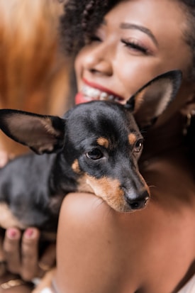 A woman is holding a small dog with black and tan fur, smiling joyfully. Her features are partially visible, with curly hair and a bright red lipstick emphasizing her smile. The intimacy and affection between the woman and the dog is apparent, creating a warm atmosphere.