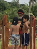 Three young children are interacting on a wooden structure in a tropical setting. The child on the left wears a baseball cap and striped shorts, while the child in the middle is wearing a black t-shirt with a design, red shorts, and a face mask. The child on the right wears blue athletic shorts. They are surrounded by lush green palm trees and foliage.
