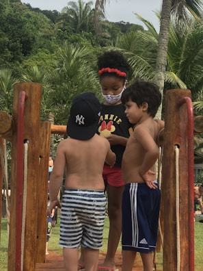 Three young children are interacting on a wooden structure in a tropical setting. The child on the left wears a baseball cap and striped shorts, while the child in the middle is wearing a black t-shirt with a design, red shorts, and a face mask. The child on the right wears blue athletic shorts. They are surrounded by lush green palm trees and foliage.