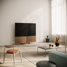 A modern living room with a large flat-screen television mounted on a stand. The room features minimalist furniture, including a wooden chair with a cushioned seat, a low coffee table with a few decorative items and a magazine, and a small vase on the floor. The floor is covered with a light-colored rug, and the room is well-lit by natural light coming through sheer curtains.