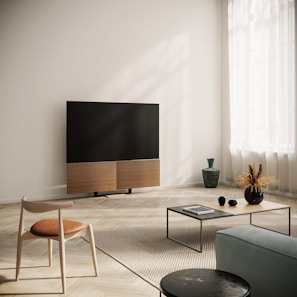A modern living room with a large flat-screen television mounted on a stand. The room features minimalist furniture, including a wooden chair with a cushioned seat, a low coffee table with a few decorative items and a magazine, and a small vase on the floor. The floor is covered with a light-colored rug, and the room is well-lit by natural light coming through sheer curtains.