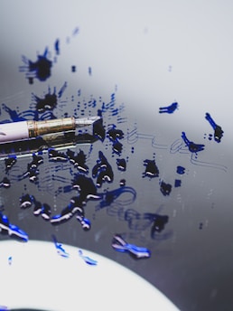 A close-up view of a fountain pen lying on a reflective surface with blue ink splattered around it. The ink forms abstract smudges and patterns, creating a sense of artistic chaos. The pen itself features a metallic gold tip and a sleek body, partially visible in the frame.
