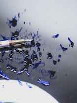 A close-up view of a fountain pen lying on a reflective surface with blue ink splattered around it. The ink forms abstract smudges and patterns, creating a sense of artistic chaos. The pen itself features a metallic gold tip and a sleek body, partially visible in the frame.