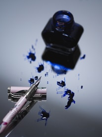 A fountain pen lies on a reflective surface next to an open ink bottle. Blue ink is splattered across the surface, creating a vibrant contrast against the smooth, mirrored background. The pen is elegantly designed, with a delicate floral pattern on its body.