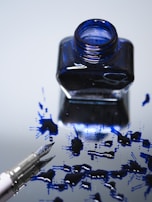 An open ink bottle surrounded by splatters of dark blue ink. Adjacent to the bottle, a fountain pen with a pointed nib rests on the surface. The scene creates a sense of creativity and artistic expression.