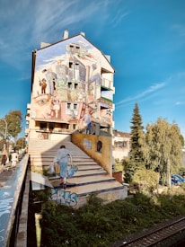A mural painted on a building facade features an imaginative scene with urban elements like buildings, staircases, and people. The artwork includes a person walking up painted stairs, seamlessly blending with the real environment. Trees and parked cars are visible in the surrounding area, contributing to the vibrant and lively atmosphere.