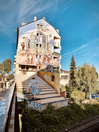 A mural painted on a building facade features an imaginative scene with urban elements like buildings, staircases, and people. The artwork includes a person walking up painted stairs, seamlessly blending with the real environment. Trees and parked cars are visible in the surrounding area, contributing to the vibrant and lively atmosphere.