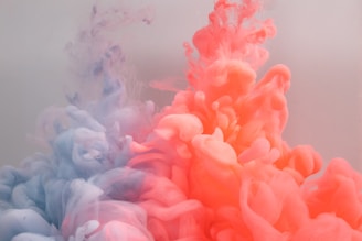 Vibrant clouds of colored ink spread and swirl through water, creating a dynamic and fluid pattern. The left side features soft purple tones transitioning into pale hues, while the right side displays a vivid coral and orange blend. The interaction of these colors forms an ethereal and abstract visual effect.