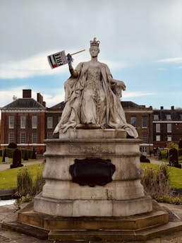 A large stone statue of a regal figure wearing a crown and long flowing robes, holding a sign in one raised hand. The statue is situated in a well-maintained garden with neatly trimmed hedges and a historic brick building in the background under a cloudy sky.