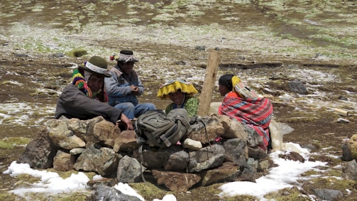 Four people wearing traditional Andean clothing are sitting on the ground in a mountainous area. They are surrounded by a circle of rocks, possibly for protection against the wind. The ground is covered with patches of snow and grass.