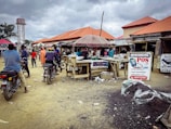 A busy outdoor market scene with several people interacting around wooden stalls. Motorbikes are parked nearby, and some individuals are seated on them. There is a large sign displaying POS services, including cash deposit and withdrawal options. The sky is overcast, and the ground is littered with debris.