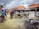 A busy outdoor market scene with several people interacting around wooden stalls. Motorbikes are parked nearby, and some individuals are seated on them. There is a large sign displaying POS services, including cash deposit and withdrawal options. The sky is overcast, and the ground is littered with debris.