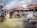 A busy outdoor market scene with several people interacting around wooden stalls. Motorbikes are parked nearby, and some individuals are seated on them. There is a large sign displaying POS services, including cash deposit and withdrawal options. The sky is overcast, and the ground is littered with debris.
