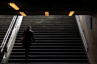 A person ascends a dimly lit stairway towards a brighter exit, with overhead lights illuminating the path and metal railings on either side.