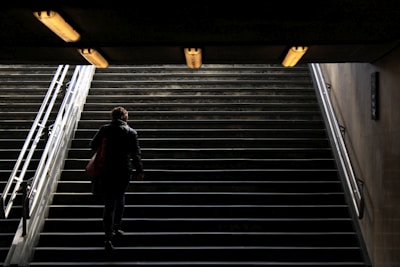 A person ascends a dimly lit stairway towards a brighter exit, with overhead lights illuminating the path and metal railings on either side.