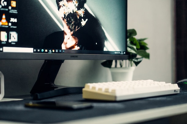 A computer setup featuring a Samsung monitor displaying a desktop with various icons. The wallpaper shows a close-up of a person holding a bouquet of flowers that obscures their face. In front of the monitor is a white keyboard. In the background, a small plant in a white pot adds a touch of greenery to the scene.