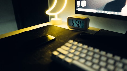 A dimly lit workspace featuring a digital alarm clock showing the time 6:50, a crescent-shaped lamp emitting a soft yellow glow, a computer monitor partly visible with a desktop screen, a smartphone placed face down on the desk, and a keyboard in the foreground.