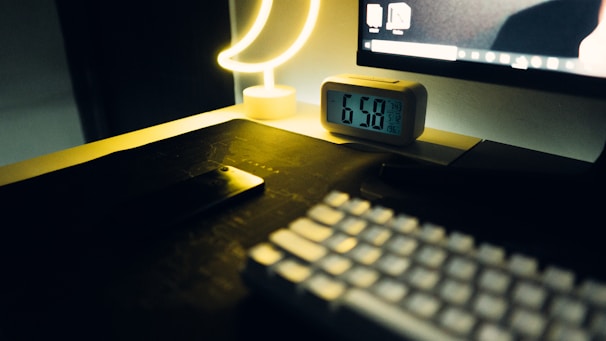 A dimly lit workspace featuring a digital alarm clock showing the time 6:50, a crescent-shaped lamp emitting a soft yellow glow, a computer monitor partly visible with a desktop screen, a smartphone placed face down on the desk, and a keyboard in the foreground.