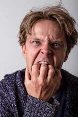A person with messy, light brown hair is looking directly at the camera with their hand covering their mouth, appearing to bite their nails. They have an expression that suggests anxiety or stress, with furrowed brows and an intense gaze. The background is plain and neutral.