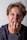 A person with messy, light brown hair is looking directly at the camera with their hand covering their mouth, appearing to bite their nails. They have an expression that suggests anxiety or stress, with furrowed brows and an intense gaze. The background is plain and neutral.