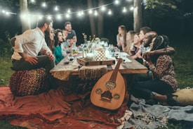 A group of people sit around a low outdoor table enjoying an evening together. They are surrounded by soft lighting from hanging string lights and lanterns illuminating a cozy setting. The table is decorated with drinks and lanterns, and one person plays a stringed instrument while others are engaged in conversation.