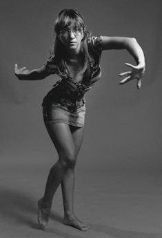 A person stands in a dynamic pose, suggesting movement or dance. They are wearing glasses, a patterned top, and a denim skirt. The backdrop is a plain, neutral tone, and the image is in black and white, focusing on the subject's expression and posture.