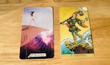 Two tarot cards are placed side by side on a wooden surface. The card on the left is modern with a woman in a short white dress walking up sand dunes, surrounded by a cosmic background with rays of light. The card on the right is traditional with a figure in colorful attire stepping near a cliff, holding a small bundle and a white dog at his side under a bright sun.
