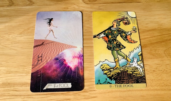 Two tarot cards are placed side by side on a wooden surface. The card on the left is modern with a woman in a short white dress walking up sand dunes, surrounded by a cosmic background with rays of light. The card on the right is traditional with a figure in colorful attire stepping near a cliff, holding a small bundle and a white dog at his side under a bright sun.