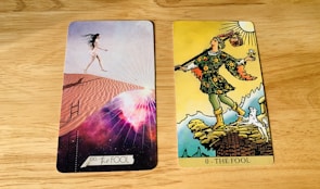 Two tarot cards are placed side by side on a wooden surface. The card on the left is modern with a woman in a short white dress walking up sand dunes, surrounded by a cosmic background with rays of light. The card on the right is traditional with a figure in colorful attire stepping near a cliff, holding a small bundle and a white dog at his side under a bright sun.