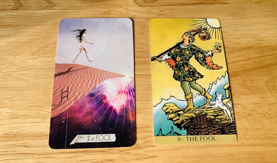 Two tarot cards are placed side by side on a wooden surface. The card on the left is modern with a woman in a short white dress walking up sand dunes, surrounded by a cosmic background with rays of light. The card on the right is traditional with a figure in colorful attire stepping near a cliff, holding a small bundle and a white dog at his side under a bright sun.