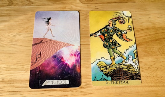 Two tarot cards are placed side by side on a wooden surface. The card on the left is modern with a woman in a short white dress walking up sand dunes, surrounded by a cosmic background with rays of light. The card on the right is traditional with a figure in colorful attire stepping near a cliff, holding a small bundle and a white dog at his side under a bright sun.