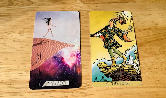Two tarot cards are placed side by side on a wooden surface. The card on the left is modern with a woman in a short white dress walking up sand dunes, surrounded by a cosmic background with rays of light. The card on the right is traditional with a figure in colorful attire stepping near a cliff, holding a small bundle and a white dog at his side under a bright sun.