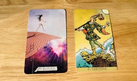 Two tarot cards are placed side by side on a wooden surface. The card on the left is modern with a woman in a short white dress walking up sand dunes, surrounded by a cosmic background with rays of light. The card on the right is traditional with a figure in colorful attire stepping near a cliff, holding a small bundle and a white dog at his side under a bright sun.