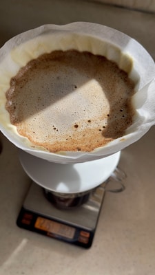 A coffee brewing setup is shown from above, featuring a paper filter containing ground coffee placed on a white coffee dripper that is set on top of a digital scale. The coffee grounds appear to be in the process of blooming.