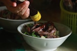 A hand is pouring a sauce over a bowl filled with sliced meat, garnished with chopped green scallions. The setting is likely a kitchen, indicated by another similar bowl in the background and a container holding utensils.