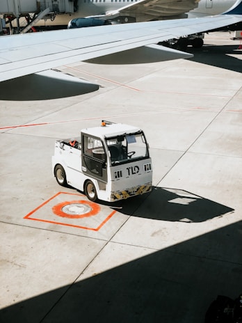 A small airport vehicle is parked on a marked spot on the tarmac. The wing of a nearby airplane casts a shadow on the concrete ground. The airplane is partially visible, including its engine and undercarriage.