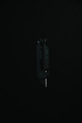 A black utility knife with a visible metal blade is placed upright against a dark background, highlighting its sleek and modern design.