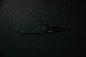 A black tactical folding knife is placed horizontally against a dark background. The knife's handle features textured grips, while its blade is partially opened, displaying a sharp edge.