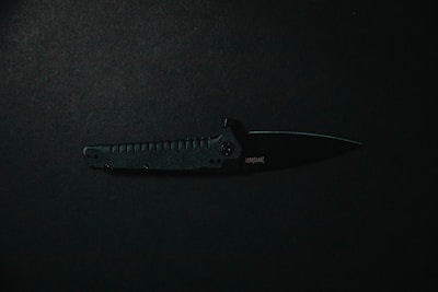 A black tactical folding knife is placed horizontally against a dark background. The knife's handle features textured grips, while its blade is partially opened, displaying a sharp edge.