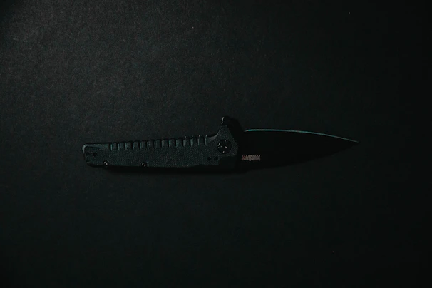 A hand gripping a tactical knife handle with textured grip, against a shadowy, industrial background.