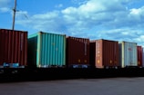 Several shipping containers are lined up in a row on a flatbed railcar under a partly cloudy sky. The containers vary in color, with blue, green, and brown tones. A utility pole with power lines is visible in the background.