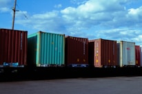 Several shipping containers are lined up in a row on a flatbed railcar under a partly cloudy sky. The containers vary in color, with blue, green, and brown tones. A utility pole with power lines is visible in the background.