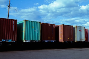 Several shipping containers are lined up in a row on a flatbed railcar under a partly cloudy sky. The containers vary in color, with blue, green, and brown tones. A utility pole with power lines is visible in the background.
