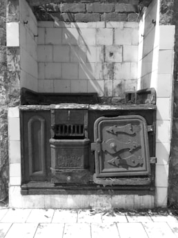 A vintage cast iron stove set against a tiled wall, featuring ornate detailing and multiple compartments. The area shows signs of age and wear, with some dirt and grime visible on the surfaces. The surrounding walls are tiled, and there is a presence of bricks and remnants of foliage at the top.