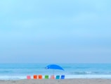 A serene beach scene featuring five colorful beach chairs in a row, including red, orange, green, and blue chairs. A large blue umbrella is set up near the chairs, providing shade. The ocean waves gently lap at the shore, and the sky is a soft blue with light clouds.
