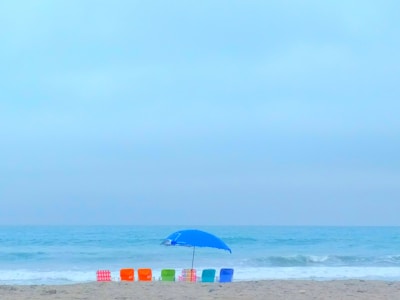 A serene beach scene featuring five colorful beach chairs in a row, including red, orange, green, and blue chairs. A large blue umbrella is set up near the chairs, providing shade. The ocean waves gently lap at the shore, and the sky is a soft blue with light clouds.