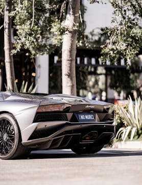A sleek, modern sports car parked on a street with a custom license plate reading 'NO MURCI'. The background features lush green foliage and a building facade partially visible.