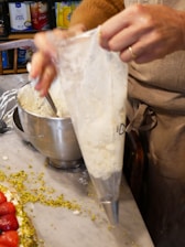 A person using a piping bag filled with cream or frosting at a kitchen countertop. There are crushed pistachios scattered around, and a partially visible cake decorated with strawberries. A mixing bowl with some batter or icing is also present in the background.