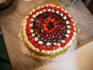 A large, round dessert arranged with layers of fresh strawberries, blueberries, raspberries, and whipped cream in a concentric pattern on a lace doily atop a marble surface. The outer rim is decorated with crushed pistachios.